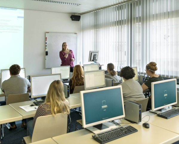 Blick vom hinteren Ende des Raums auf eine Schulungsveranstaltung. Vorne spricht eine weibliche Person und zeigt etwas auf einer Power-Point-Pr?sentation. Die Kurseteilnehmenden sitzen an Tischen vor Coumputerbildschirmen.