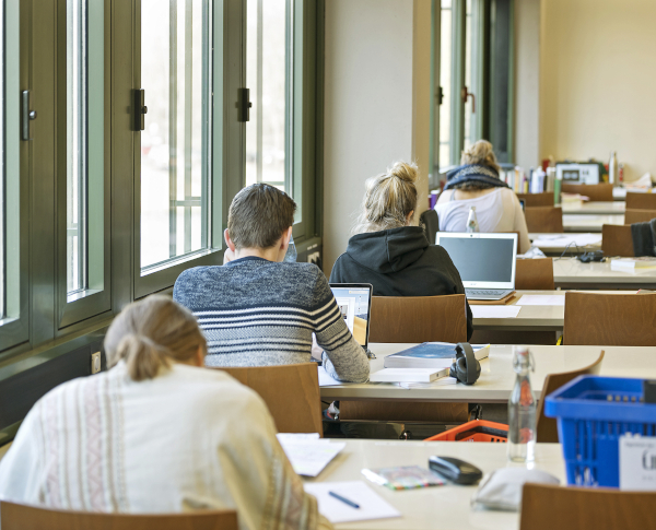 Personen sitzen in Tischreihen hintereinander. Die Personen arbeiten am Laptop und lesen in B��chern.