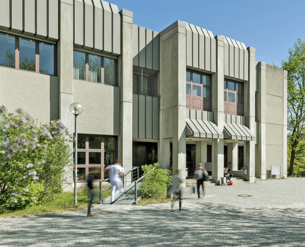 Blick von vorne auf den Eingangsbereich eines brutalistischen Beton-Geb?udes. Links und rechts sieht man gr��ne B?ume und Str?ucher. Vier Personen befinden sich auf dem Weg ins Geb?ude.