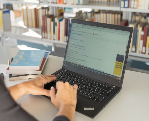 Auf einem Tisch steht ein aufgeklappter Laptop. Einer Person tippt auf der Tastatur. Man sieht nur die H?nde und Unterarme. Auf dem Tisch liegen B��cher. Im Hintergrund stehen B��cherregale.