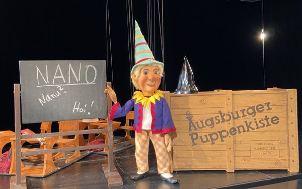 Das bekannte Kasperl aus der Augsburger Puppgenkiste steht vor einer Tafel, auf der Nano, nanu, hoi geschrieben steht. Es geht dabei um die Kinderuni mit dem Thema Expedition in die Winzigkeit.