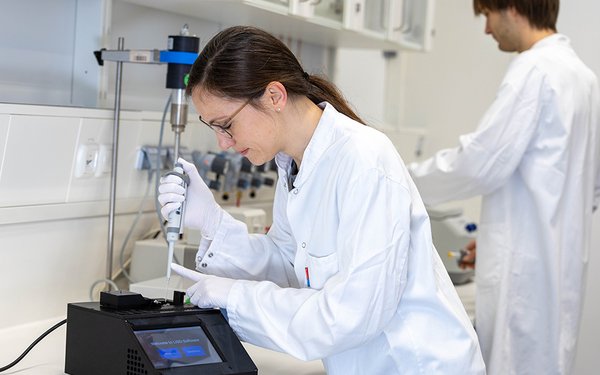 In einem Labor arbeitet eine Frau in weißem Kittel an einem schwarzen, kastenförmigen Gerät, das etwas kleiner als ein Schuhkarton ist. Auf dem Display des Geräts steht „Welcome to LISO-Software“.