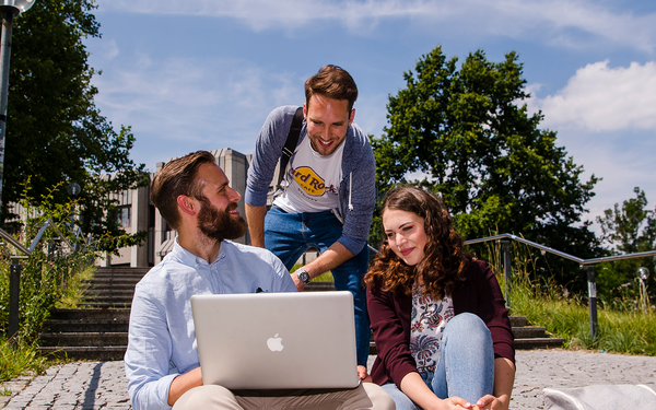 Eine Gruppe von zwei Studenten und einer Studentin sitzt auf einer Treppe und schaut gemeinsam in einen Laptop. Im Hintergrund sieht man den grünen Campus.