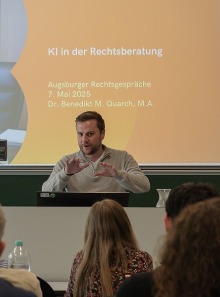 Herr Dr. Quarch hält im Rahmen der Augsburger Rechtsgespräche einen Vortrag. Im Hintergrund ist eine Powerpoint-Präsentation zu sehen.