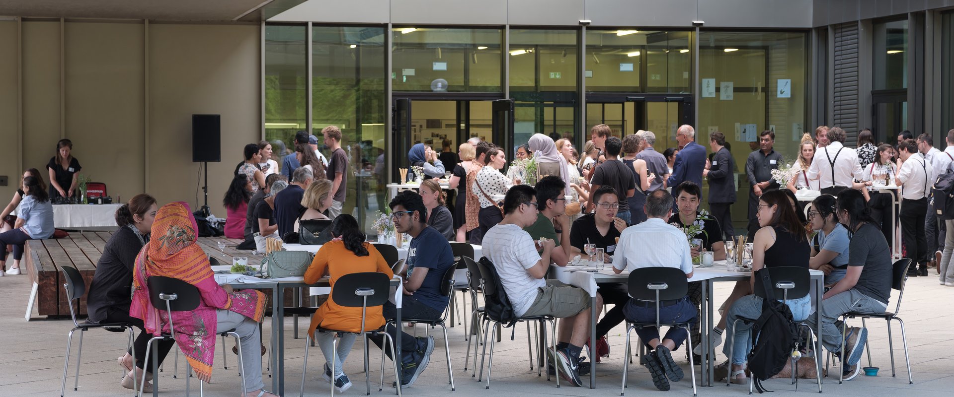 Visitors to the Summer Festival 2023; people at tables and bar tables chatting lively in the courtyard of the Building for Music and Art