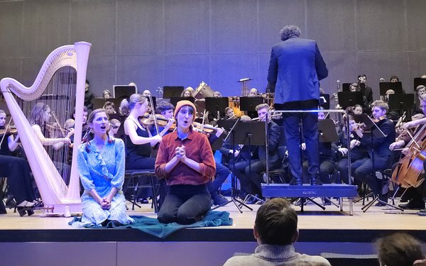 Die beiden Solistinnen knien auf der B��hne. Hinter ihnen eine Harfe und das komplette Universit?tsorchester. Mit dem R��cken zu ihnen dirigiert Dr. Christoph Teichner.
