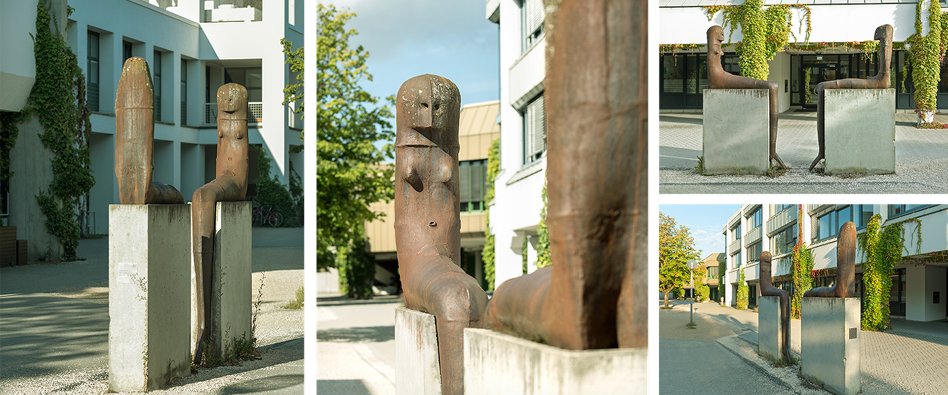 Auf dem Bild sieht man das Kunstwerk von Lothar Fischer, bei dem sich zwei Skulpturen gegenübersitzen auf Steinen.