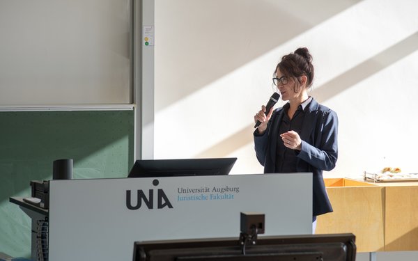 Pia Lorenz steht mit einem Mikrofon in der Hand im Hörsaal vor einer grünen Tafel und hält einen Vortrag.
