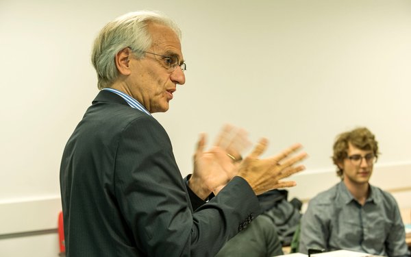 Prof. Dr. Christoph Weller ist im Profil zu sehen. Er hat graues Haar und gestikuliert in Richtung eines Publikums, vor dem er spricht. Rechts im Bild sieht man eine zuhörende männliche Person.