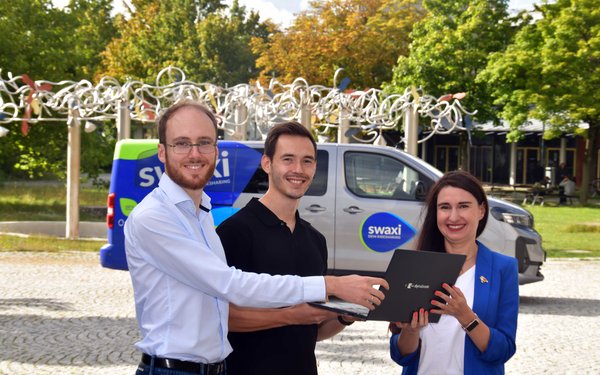 David Fleckenstein und Fabian Anzenhofer stehen gemeinsam mit Julia Boxler vor einem swaxi und lächeln in die Kamera. Sie halten gemeinsam ein Dokument in der Hand.