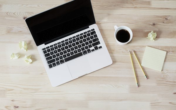 BIld auf dem ein Laptop, eine Tasse Kaffe, ein Stift und ein paar zerknüllte Post-its zu sehen sind