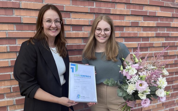 Anabela Brandao und Nina Hümmer stehen an einer roten Klinkersteinwand und lächeln in die Kamera. Hümmer trägt einen Blumenstrauß, Brandao die Urkunde.