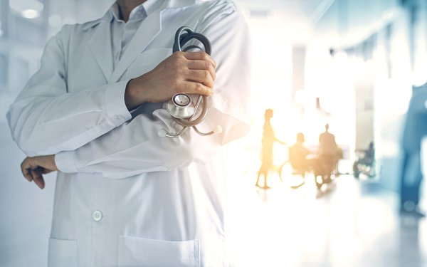 Es ist ein Arzt in einem weißen Kittel mit einem Stethoskop zu sehen. Im Hintergrund sind verschwommene Silhouetten von Menschen in einem Krankenhausflur erkennbar.