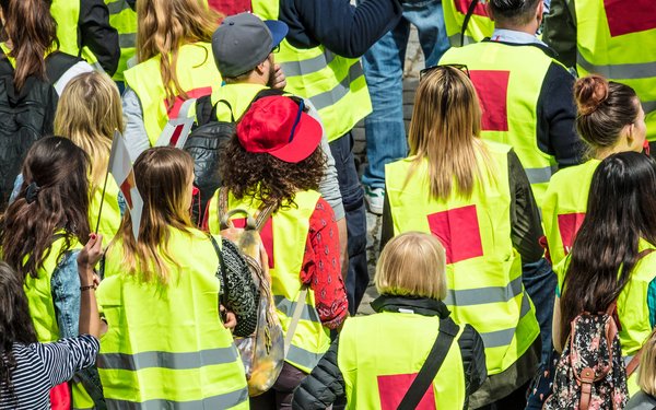 Eine Menschenversammlung von hinten. Die Personen tragen gelbe Warnwesten mit roten Aufdrucken.