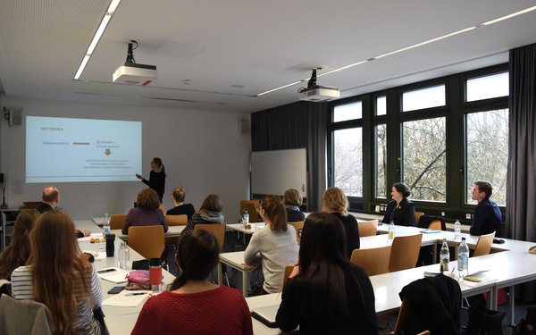 In einem Seminarraum sitzen einige Personen, die nach vorne auf eine vortragende Person blicken. Diese hat ihre Präsentation an die Wand projiziert und ist dieser erklärend zugewandt.
