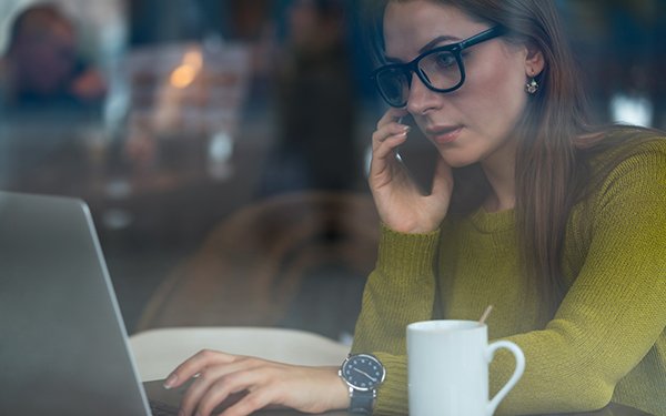 Eine brünette Frau mit einer schwarzen Brille sitzt vor einem aufgeklappten Laptop und telefoniert. Dabei blickt sie in den Laptop und neben ihr steht eine weiße Kaffeetasse mit einem Löffel. Der Hintergrund ist unscharf.