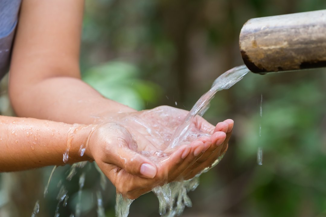 Zwei Hände sind zu einer Schale geformt und fangen Wasser auf, das aus einem braunen Rohr läuft. Im Hintergrund Waldatmosphäre.