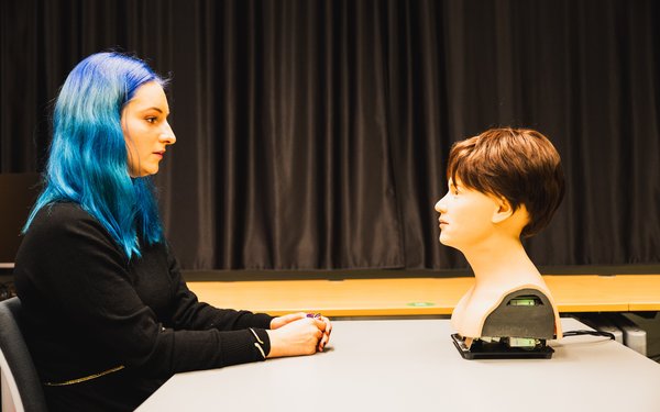 Johanna Kuch sits directly opposite the robot head, which is standing on a table.