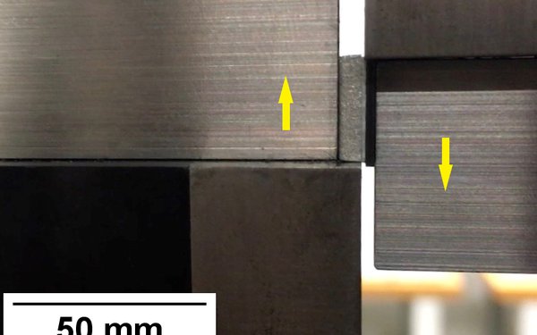 Man sieht zwei sich in gegenläufige Richtung bewegende Metallblöcke, deren Bewegungsrichtung mit Pfeilen gekennzeichnet ist.