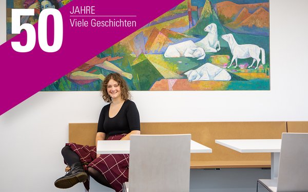 Die Studierende Sybille Lang sitzt, mit Blick in die Kamera, an einem Tisch mit einem Wandbild hinter sich.