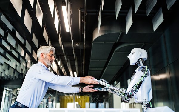 A man with a grey beard and grey hair and a robot are standig face to face in a dark hall and are holding hands.