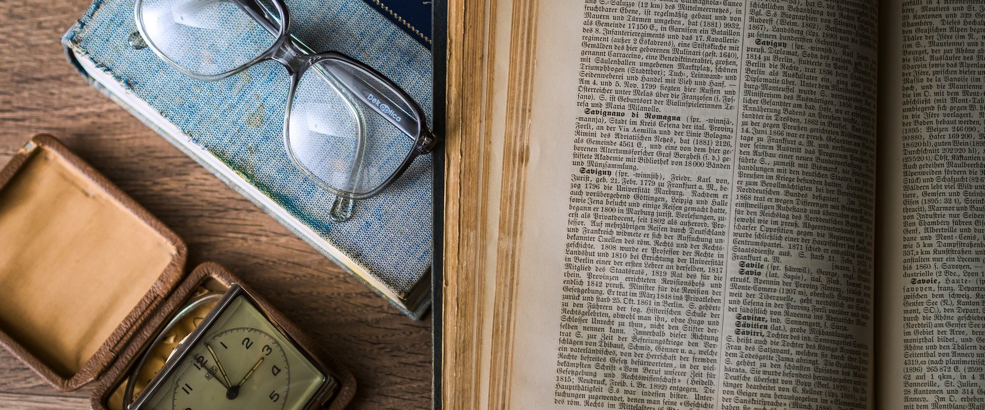 Bild zeigt ein aufgeschlagenes Buch, eine Brille, eine Uhr