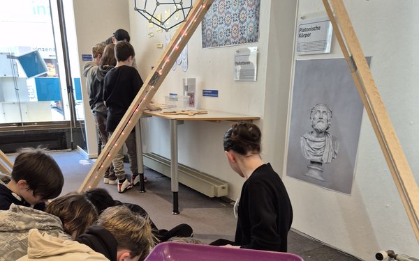 Im Vordergrund eine Schale mit roten und blauen Würfeln und einer Arbeitsanweisung, im Hintergrund beschäftigen sich Schüler mit geometrischen Figuren
