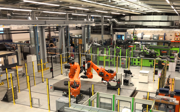 Blick von oben in den KI-Erlebnisraum "Halle 43". Grüne, gelbe und orange Gerätschaften und Roboter zwischen weißen Tischen.