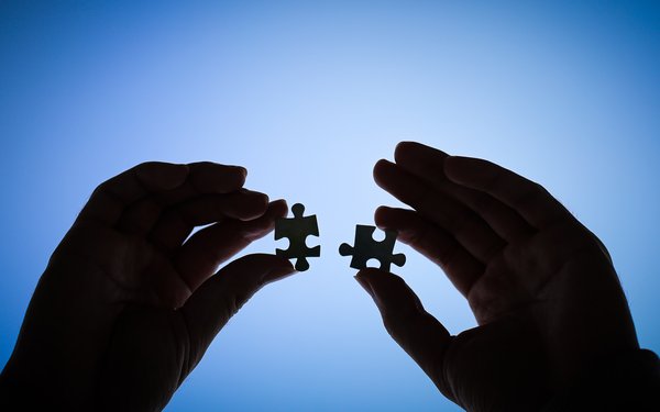 Zwei Hände halten jeweils zwei Puzzle teilen und sind nur silouttenhaft zu sehen. Die Puzzleteile sollen symbolisch für Inklusion stehen. Das Konzept ist dann das Puzzle, bei dem alle Teile perfekt zusammen passen.