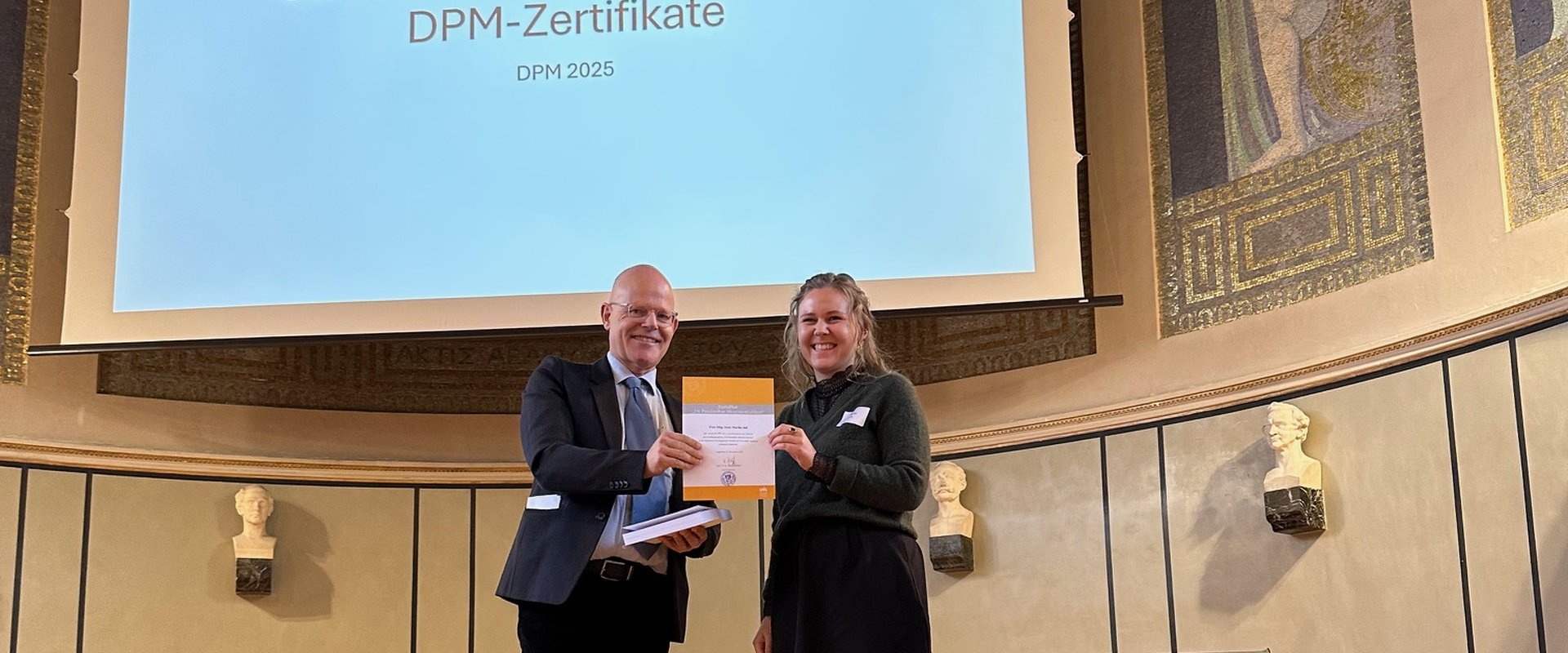 Prof. Dr. Dr. Elmar Gthoff berreicht das Zertifikat DPM an Marike Jall. Das Bild zeigt beide auf der Bhne der Gro?en Aula der LMU Mnchen.