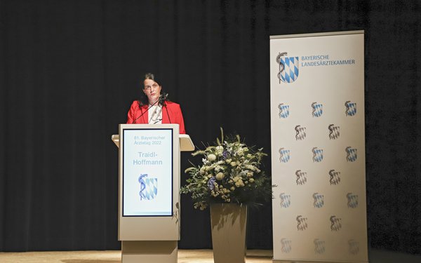 Frau Traidl-Hoffman steht im roten Blazer an einem Sprechpult. Rechts auf dem Bild ein Banner der Bayerischen Landesärztekammer und ein Blumengesteck.