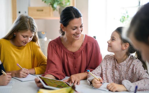 In der Mitte eine junge Lehrerin mit rotem Oberteil und dunklen langen Haaren, die am Tisch sitzt. Rechts neben ihr ein Mädchen und ein junge mit Tablet. Links neben ihr eine junge Frau in gelbem Oberteil und langen, blonden Haaren, die etwas aufschreibt.