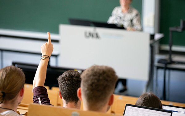 Studierende in einer Vorlesung mit Dozentin, ein Student meldet sich