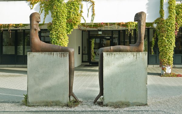 Auf dem Bild sieht man das Kunstwerk von Lothar Fischer, bei dem sich zwei Skulpturen gegenübersitzen auf Steinen.