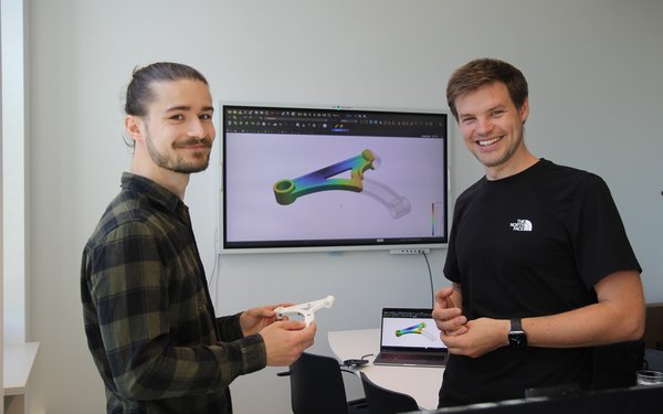 Nils Meyer (rechts) und Projekt-Mitarbeiter Julian Greif lachen in die Kamera, während sie gerade ein KI-Tool entwickeln, das die Bauteilentwicklung im Spritzguss optimiert.