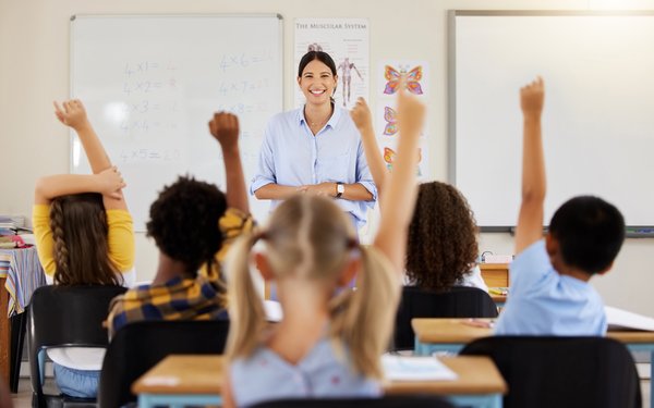 Eine junge, schwarzhaarige Lehrerin l?chelt in die Kamera. Im Vordergrund sitzen Schler an Tischen und sind von hinten zu sehen. Einige melden sich.