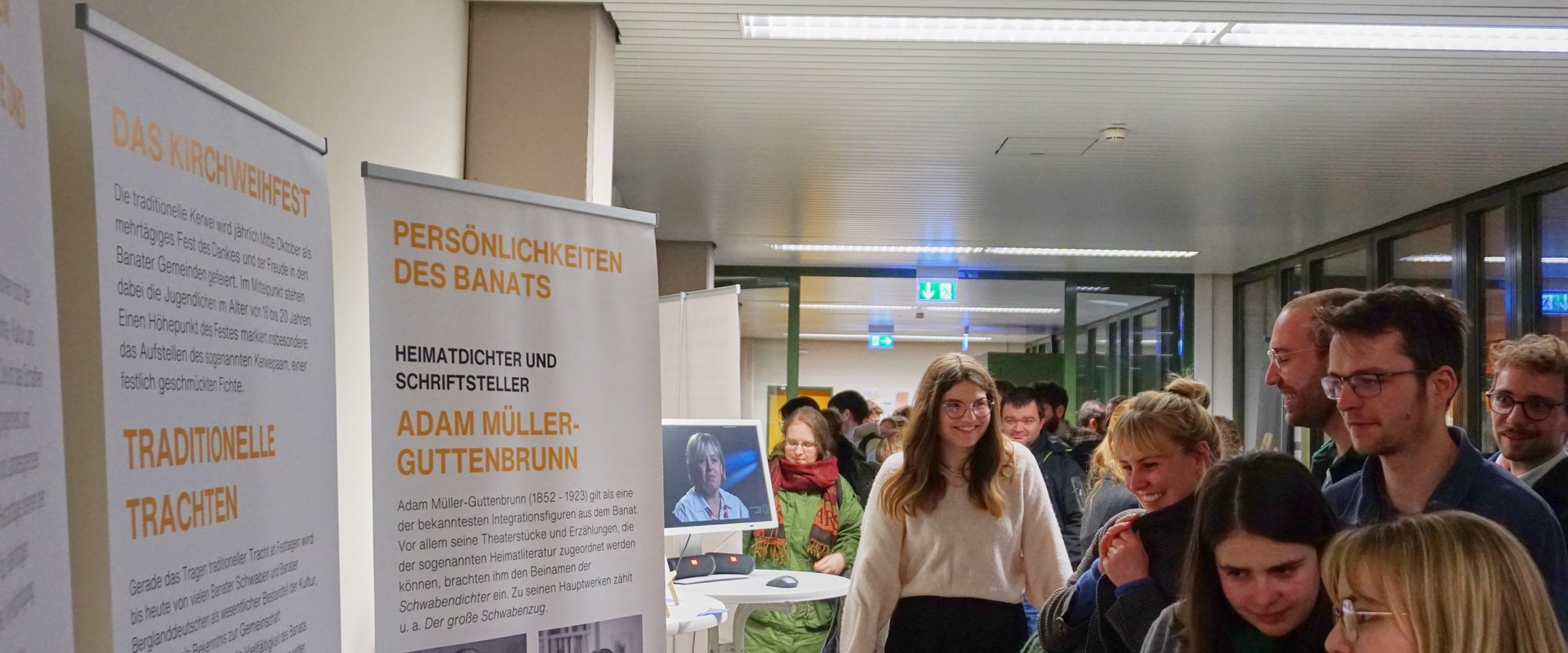 Es sind Student:innen und Besucher:innen in Interaktion zu sehen, die vor einer Stellwand stehen.