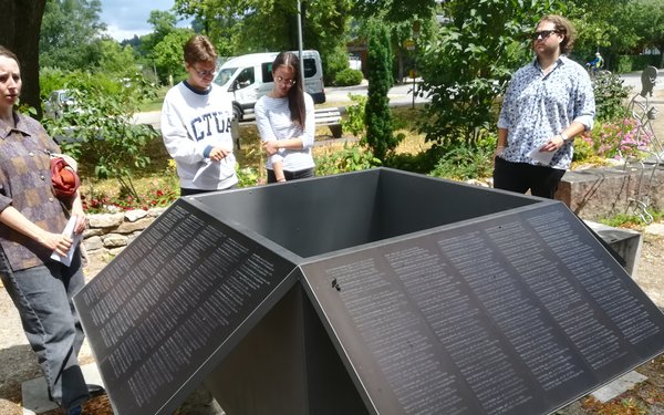 Vier Studierende stehen im grünen Gelände vor Gedenktafeln, die in einem Quadrat angeordnet sind. Die Tafeln sind schwarz und es sind viele Namen darauf sichtbar.
