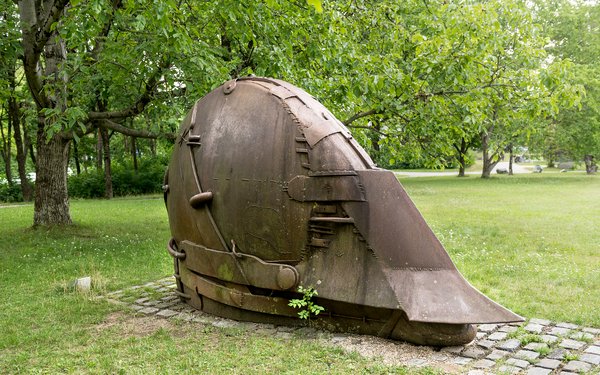 Das Bild zeigt einen metallische Skulptur in Form eines Helms in brauner Farbe inmitten einer grünen Wiese