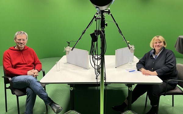 Vor gr��nem Hintergrund sitzen links Prof. Dr. Stefan K��nzell und rechts Prof. Dr. Sabine Doering-Manteuffel am Tisch und l?cheln in die Kamera. Vor ihnen h?ngen Mikrofone von der Decke