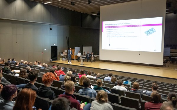 Auf einer Bühne im Musikauditorium der Uni AUgsburg steht eine Person und hält einen Vortrag.