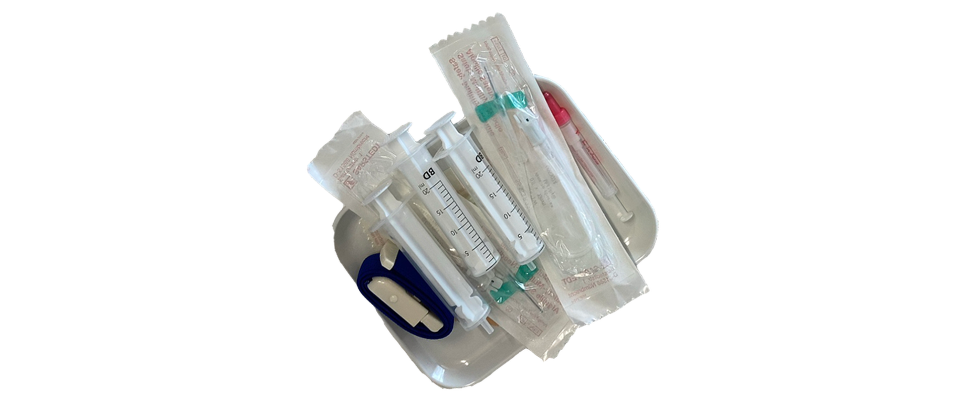 Das Blutentname Set besteht aus zwei kleinen durchsichtigen Spritzen ohne Nadel und zwei verpackten sterilen Kanülen und einem Venenstauer, um die Blutentnahme zu erleichern. Alles liegt in einer kleinen Schale.