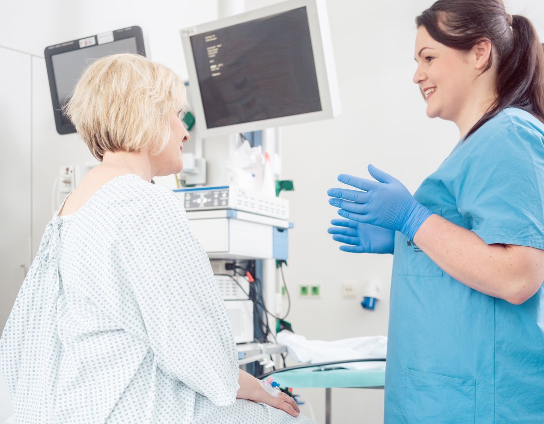 Eine Frau sitzt im Krankenhaushemd auf einer Liege und redet mit einer Vertreterin des medizinischen Personals.