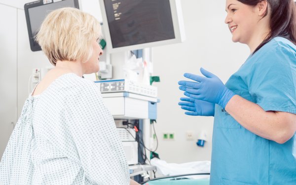 Eine Frau sitzt im Krankenhaushemd auf einer Liege und redet mit einer Vertreterin des medizinischen Personals.