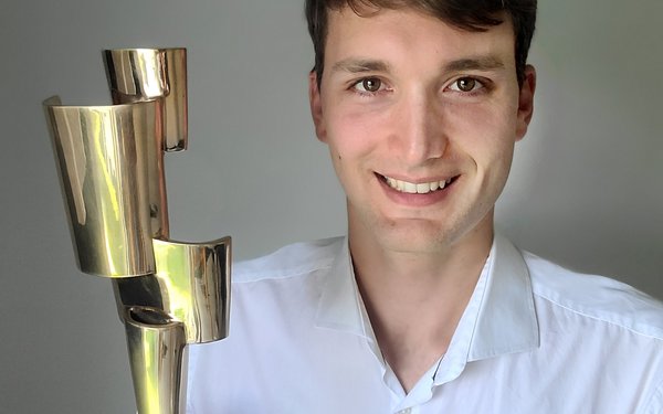 Dr. Moritz Hauck lacht in die Kamera und hat in der rechten Hand den länglichen, goldenen Kulturpreis