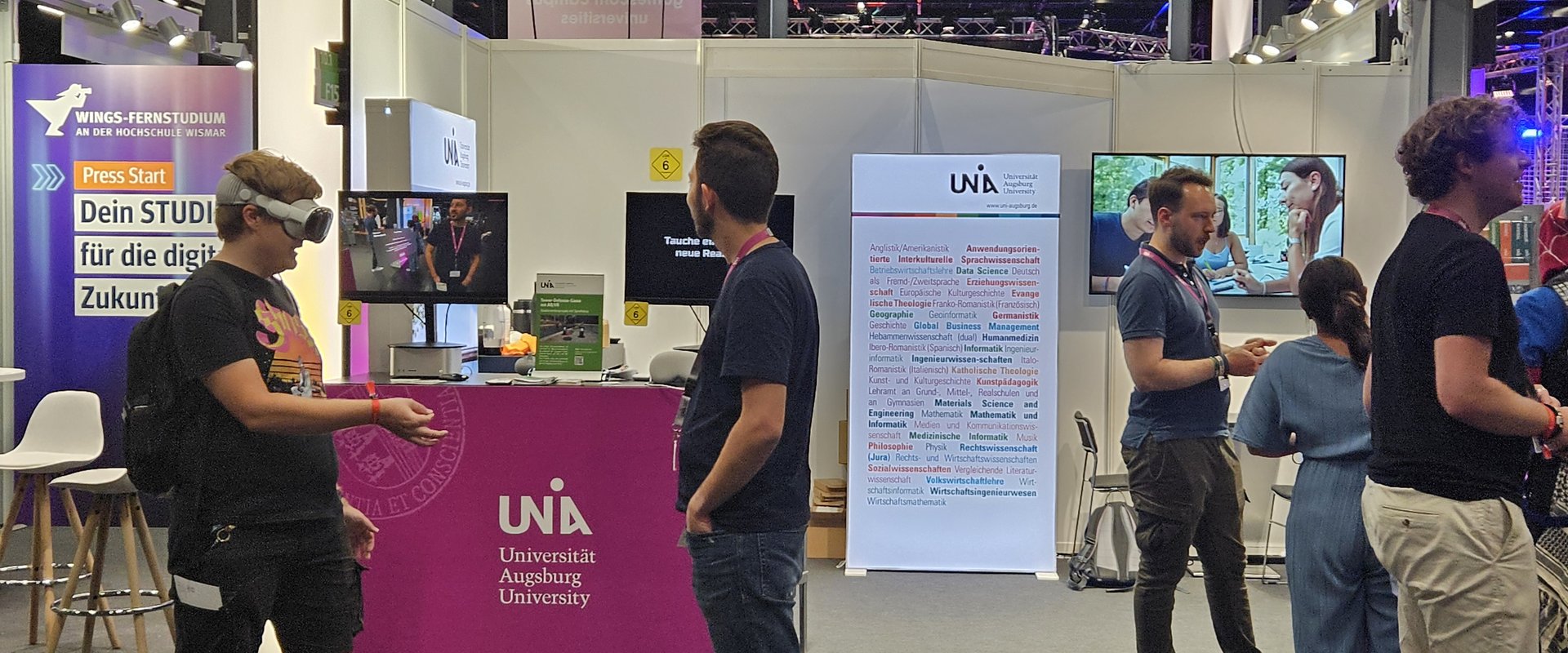Several people are standing at the exhibition stand. On the left is someone wearing VR glasses, on the right are several people engaged in conversation.