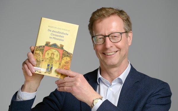 Prof. Dr. Martin Kaufhold lächelt in die Kamera und hält ein Exemplar seines neu erschienenen Buches "Die abendländische Christenheit im Mittelalter" hoch.