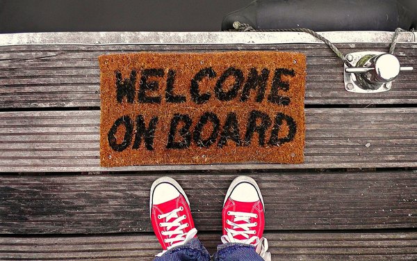 Füße in Turnschuhen vor Fußmatte mit der Aufschrift "Welcome on Board"
