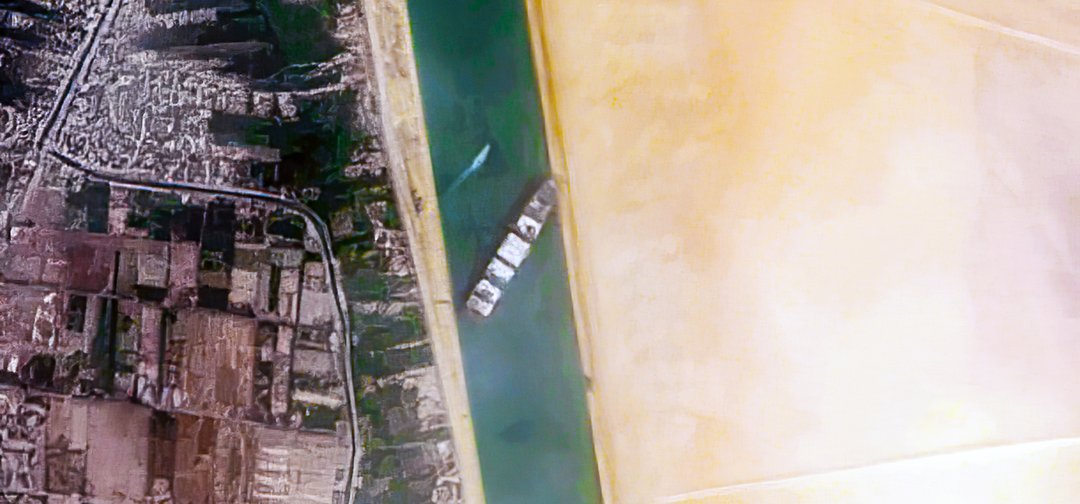 Das Bild wurde von oben aufgenommen und zeigt den Suez-Kanal, welcher von einem Frachtschiff blockiert wird.