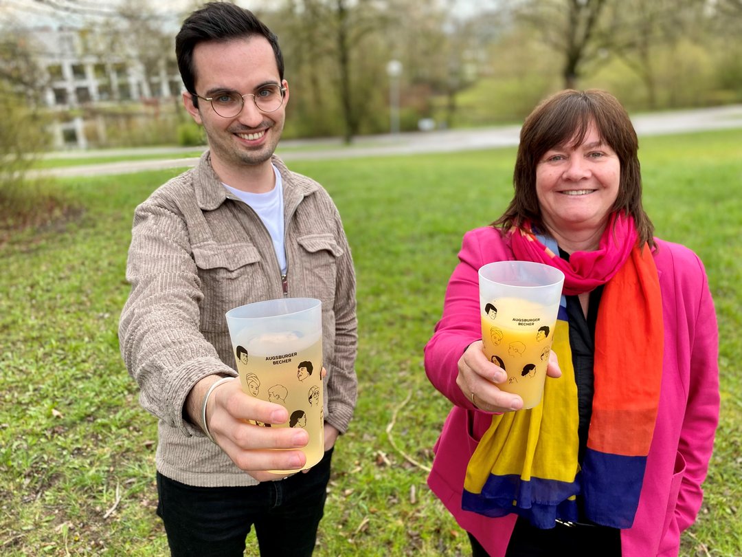 Herr Assies und Frau Thorenz auf dem Campus, in der Hand halten sie jeweils einen "Augsburger Becher"